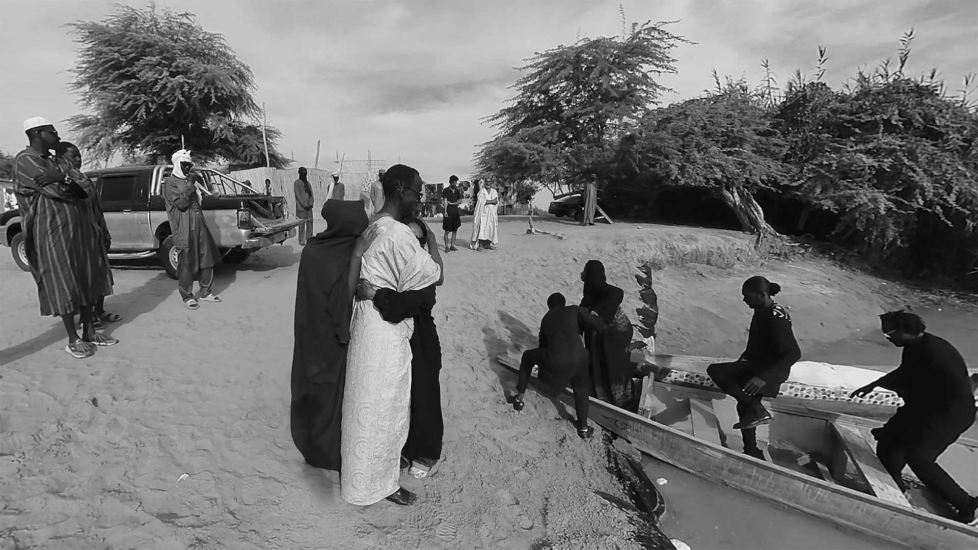 Sur une berge de sable, deux femmes s'enlacent pendant que des proches chargent une pirogue