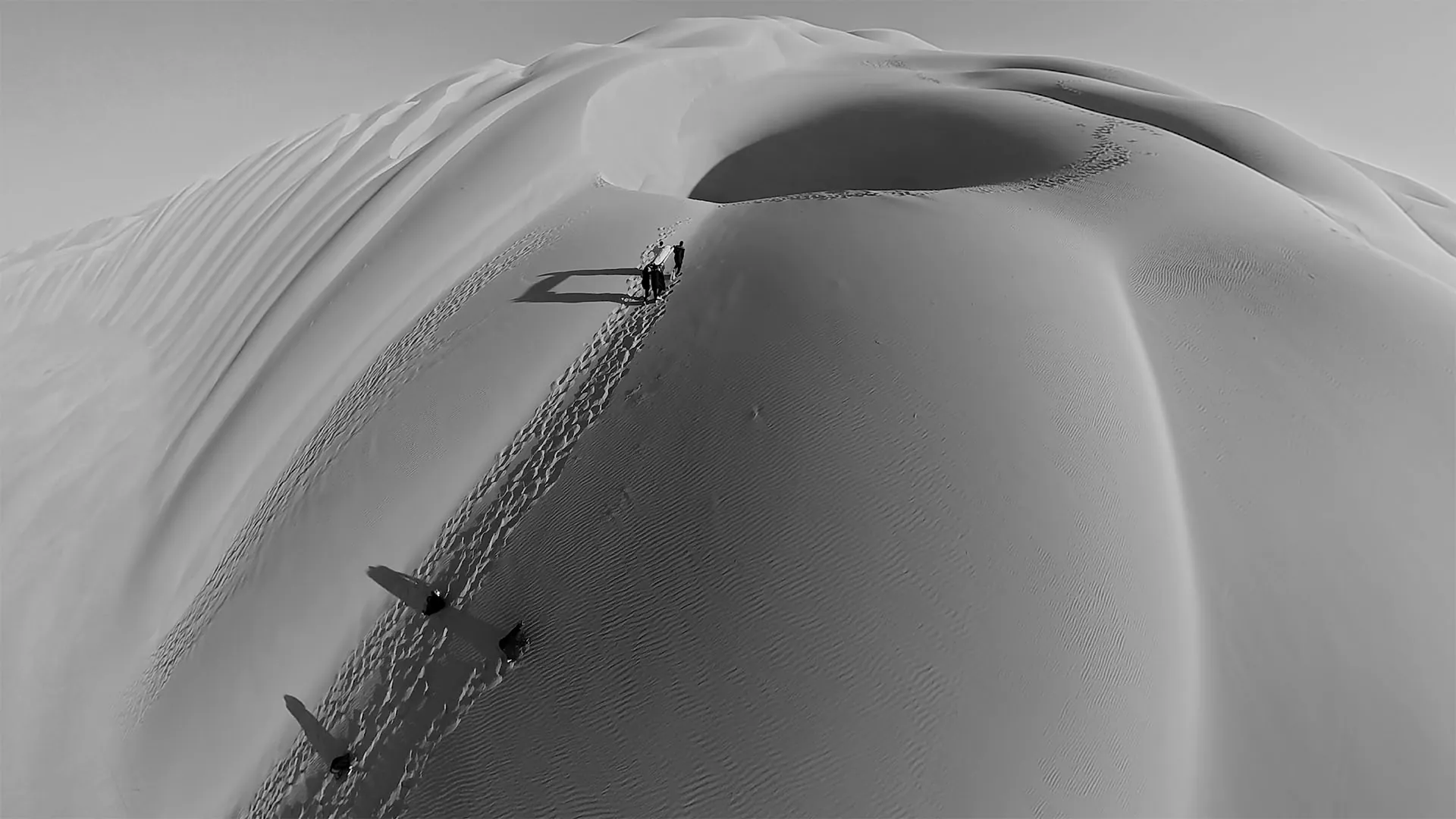 Vue aérienne : quelques silhouettes gravissent la crête d'une immense dune, laissant une trace de pas